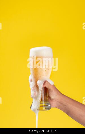 il braccio dell'uomo contiene un bicchiere di vetro con birra all'interno, schiuma bianca e fondo giallo solido, la schiuma cade attraverso il bicchiere e sul braccio Foto Stock