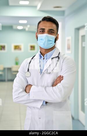 Maschio medico indiano felice indossare facemask in piedi in clinica ospedale. Verticale Foto Stock