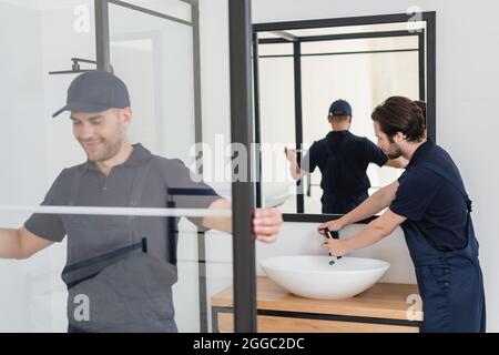 sorridente handyman misurare cabina doccia vicino idraulico lavoro con lavandino in bagno Foto Stock