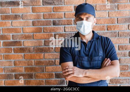 uomo in maschera medica in piedi con braccia incrociate vicino muro di mattoni Foto Stock