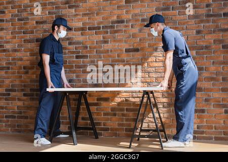 lavoratori in tute e maschere mediche che tengono legno compensato vicino muro di mattoni Foto Stock