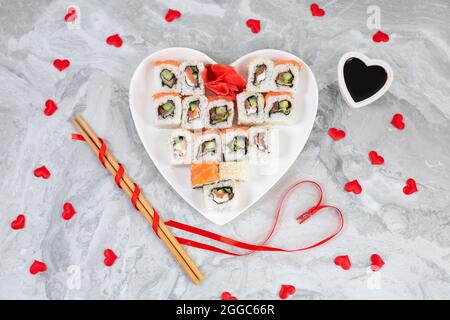 Rotoli di sushi con salmone su piatto a forma di cuore bianco e bastoni di  sushi in legno tra cuori di confetti rossi su sfondo di marmo grigio.  Concetto di cibo per