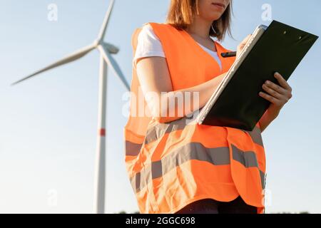 La donna nel giubbotto arancione prende appunti sulla manutenzione delle turbine eoliche sullo sfondo del mulino a vento alla luce del sole Foto Stock