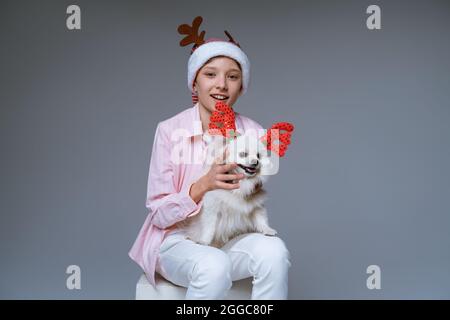 Il ragazzo allegro in cappello con le corna di cervi tiene il cane tra le braccia, che è anche con le corna di cervi su sfondo chiaro. Divertente foto concetto di natale. Ragazzo caucasico babbo natale Foto Stock