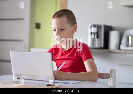 Boy 6-10 completa le lezioni scolastiche e i compiti online in cucina, guardando il computer tablet. Il concetto di istruzione a distanza. Rimani a casa. Foto Stock