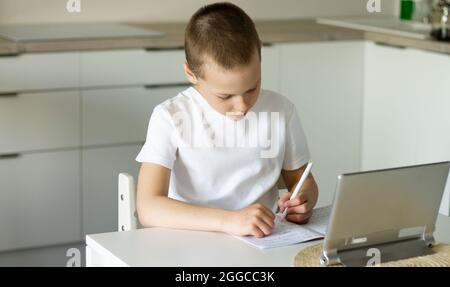 Boy 6-10 completa le lezioni scolastiche e i compiti online in cucina, guardando il computer tablet. Il concetto di istruzione a distanza. Rimani a casa. Foto Stock