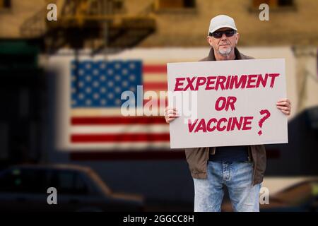 Uomo dispiaciuto con cappuccio, jeans blu e occhiali da sole che tengono un ESPERIMENTO SENZA segno di vaccino COVID O VACCINO con stelle e strisce bandiera su un muro nel Foto Stock