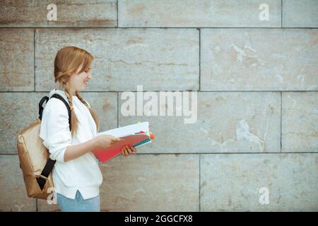 felice pupilla moderna in felpa bianca con libro da lavoro e zaino contro muro all'aperto in città. Foto Stock