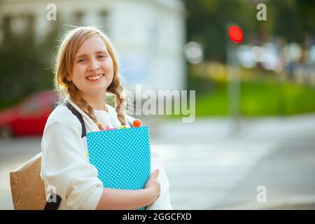 Ritratto di ragazza elegante sorridente in felpa bianca con libro degli esercizi e zaino che attraversa il crosswalk e va a scuola all'aperto in città. Foto Stock