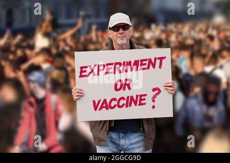 Uomo dispiaciuto con cappuccio, jeans blu e occhiali da sole che tengono un esperimento di segno di vaccino contro COVID O VACCINO con una folla di persone in background. Anti-vaxx Foto Stock