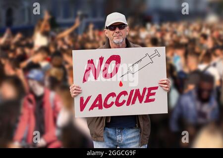 Uomo dispiaciuto con cappuccio, jeans blu e occhiali da sole che tengono un SEGNO DI VACCINO CONTRO COVID con folla di persone in background. Supporto della MOV. Anti-vaccinazione Foto Stock