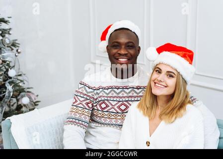 Una giovane coppia multietnica a santa cappelli sono seduti su un divano in un luminoso interno accanto ad un albero. Uomo e donna guardando la macchina fotografica, sorridendo. Foto Stock