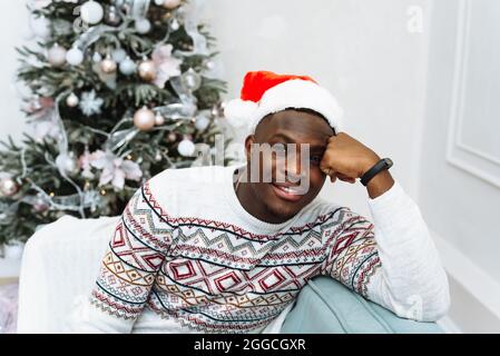 Ritratto di un bel giovane uomo nero sorridente e guardando la macchina fotografica che porta un cappello di Natale su un divano con un albero di Natale sullo sfondo. Foto Stock