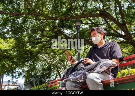 uomo con una maschera per il viso seduta su una panca da parco utilizzando il suo smartphone Foto Stock