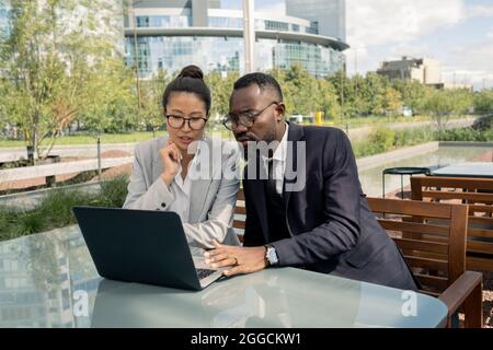 Due giovani colleghi in abbigliamento formale e occhiali seduti al tavolo davanti al computer portatile all'aperto Foto Stock