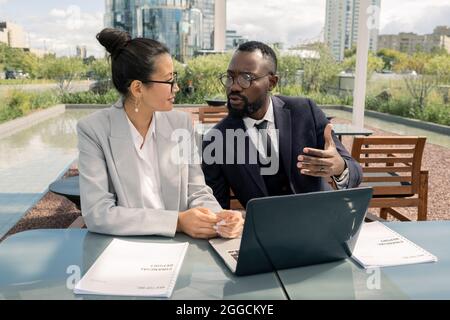 Due giovani colleghi interculturali discutono di punti di presentazione davanti al notebook all'aperto Foto Stock