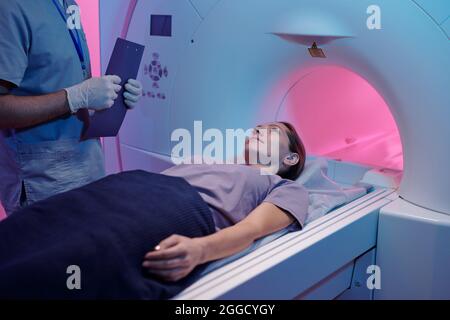 Giovane donna sdraiata sul lungo tavolo della macchina di scansione mri mentre il medico sta accanto a lei Foto Stock