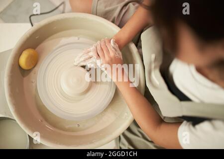 Vista dall'alto di un vasaio femminile anonimo che scolpisce il materiale argilloso sulla ruota rotante durante il lavoro in officina professionale Foto Stock