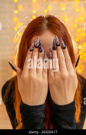 Donna della strega dello zenzero di Halloween con le unghie lunghe spaventose. Garland sullo sfondo. Le mani delle donne coprono il suo viso. Foto Stock