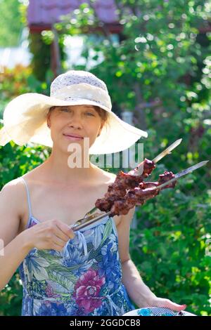 Una giovane donna graziosa in un cappello di paglia nel sole luminoso in una giornata estiva tiene un kebab fresco shish fragrante nelle sue mani. Primo piano. Verticale Foto Stock