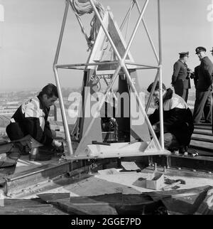 I lavoratori di Laing sul tetto della cattedrale di Coventry che imbullonano la guglia di bronzo da 80 piedi ad una piastra quadrata di 1 iarda, con gli ufficiali della RAF in piedi sullo sfondo. Le persone sul tetto, tra cui il personale dell'Aeronautica reale e il personale di Laing, facevano parte dell'operazione "Operation Rich Man". Questo ha coinvolto un elicottero belvedere RAF che si è issato per posizionare la guglia di bronzo da 80 piedi sulla nuova cattedrale. La parte dell'operazione di abbassamento della traversa di 1/2 tonnellate che si trova sulla sommità della guglia doveva essere posticipata a causa delle condizioni di vento. Foto Stock