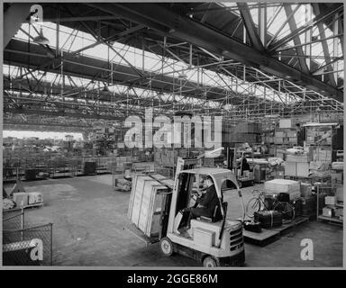 Guardando attraverso l'interno del capannone di trasporto e pacchi presso il British Road Services Depot, mostra un conducente di carrelli elevatori che sposta le casse in primo piano. Il capannone di trasporto e pacchi fu costruito da Laing alla fine degli anni '50 per British Road Services (in seguito National Freight Corporation). Aveva un'ampia area del pavimento senza ostacoli resa possibile dall'uso di massicce travi in traliccio che sostengono la struttura. Foto Stock