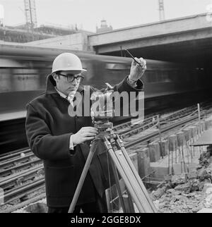 Un topografo laing, che utilizzava uno strumento teodolitico, si trovava di fronte ad una linea ferroviaria nel cantiere dello sviluppo barbicano. Nel 1962 sono iniziati i lavori per la costruzione dello sviluppo barbarico di Londra&#x2019;s. La costruzione di questa complessa megastruttura è stata progettata per essere realizzata in sei fasi, con diversi appaltatori che lavorano sul sito. Nel 1964 John Laing and Son Ltd ha vinto la gara per la fase III, e hanno finito Speed House nel 1968. La Società è stata anche responsabile della costruzione del Barbican Arts Center (fase V), iniziato nei primi anni '70 e completato nel 1981. Foto Stock