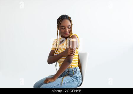 Allegra donna nera dai capelli lunghi millennial in casual seduta su sedia su sfondo bianco, guardando l'intonaco medico sulla spalla e sorridendo, Foto Stock