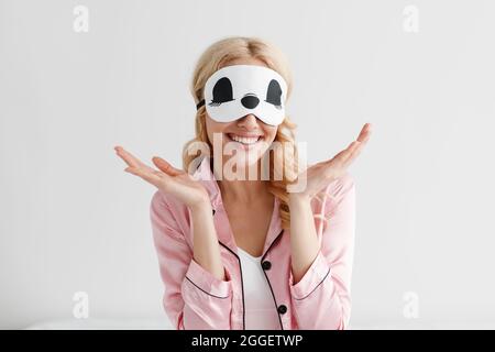 Divertente signora godendo tempo libero e buon mattino, soggiorno a casa, convivid-19. Soddisfatto bella giovane donna caucasica in pajama e la maschera di sonno si diffonde a Foto Stock