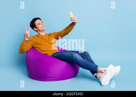 Foto a tutta lunghezza di divertente brunet millennial guy sit on bag do selfie v-sign indossare jeans maglione isolato su sfondo blu Foto Stock