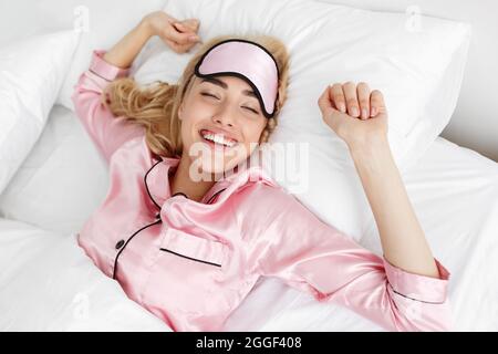 Felice donna giovane e rilassata con maschera di sonno svegliata in camera da letto accogliente, mani stretching, sensazione di energia. Sorridente bionda caucasica millenaria godendo Foto Stock