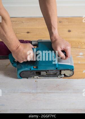 Primo piano delle mani maschili caucasiche azionamento di una levigatrice a nastro per rimuovere la vernice bianca da un pavimento di legno. Foto Stock