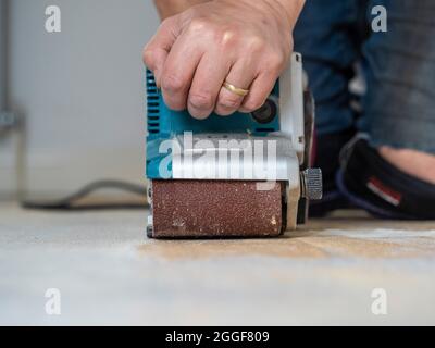 Primo piano di mano maschio caucasica azionamento di una levigatrice a nastro per rimuovere la vernice bianca da un pavimento di legno. Foto Stock