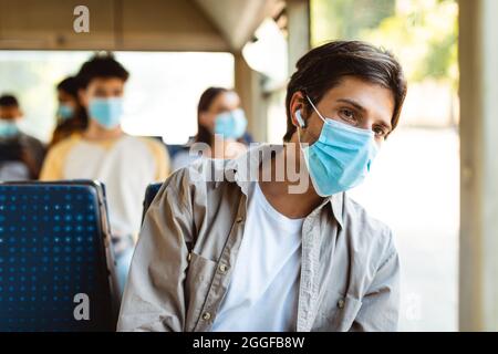 Stile di vita moderno. Casual uomo giovane indossare maschera chirurgica viso e auricolari wireless viaggiando in tram in città, seduto vicino alla finestra e guardando fuori, lis Foto Stock