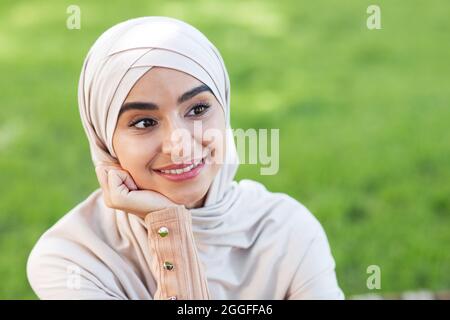 Ritratto di sorridente e pensiva bella giovane musulmana araba in hijab, guardando lo spazio vuoto nel parco su sfondo verde erba, primo piano. Bellissima pazza Foto Stock