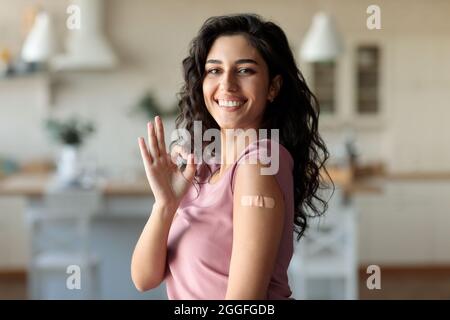 Sorridendo giovane donna caucasica che mostra un gesto corretto dopo l'iniezione di vaccino covid-19, avendo la fascia di aiuto sul suo braccio, raccomandando l'immunizzazione coronavio a Foto Stock