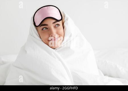 Emozioni facciali positive, buon mattino dopo un sonno ristoratore. Soddisfatto sognante divertente giovane donna caucasica in maschera di sonno avvolto su coperta in camera da letto e. Foto Stock