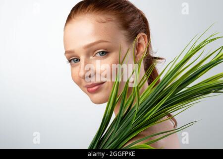 Pretty Young Lady Holding Green Plant near Face Smiling to Camera Standing Over Grey background in Studio. Cura del viso, cura del corpo e Spa. Naturale Foto Stock