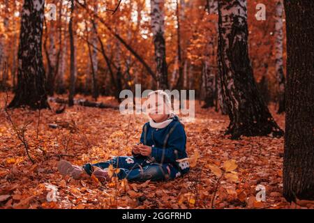 intera lunghezza concetto di infanzia felice. sorridente bella bambina in cappotto di jeans blu e jeans sedersi su terra coperta di foglie rosse cadute in autunno Foto Stock