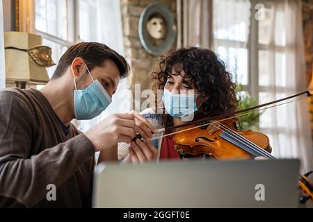 La donna felice suona il violino sotto le istruzioni dell'insegnante di musica in maschera durante il coronavirus a casa Foto Stock