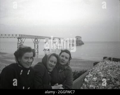 Anni '50, storica, tre giovani donne sulla spiaggia di fronte alla passerella in legno e vecchia stazione di scialuppa di salvataggio a Selsey, West Sussex, Inghilterra, Regno Unito. Un servizio di scialuppa di salvataggio è stato istituito a Selsey nel 1861. Nel 1927 per ospitare il nuovo motonave della stazione, la casa barca è stata costruita e questa struttura è visibile nella foto. Questa stazione di scivoli fu nuovamente ricostruita alla fine degli anni '50, ma fu abbassata nel 2017. Foto Stock