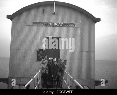 1950, storico, persone in piedi sulla passerella di legno di fronte alla vecchia stazione di scialuppa di salvataggio a Selsey, West Sussex, Inghilterra, Regno Unito. Un servizio di scialuppa di salvataggio è stato istituito a Selsey nel 1861. Nel 1927 per ospitare il nuovo motonave della stazione, la casa barca è stata costruita e questa struttura è visibile nella foto. Questa stazione di scivoli fu nuovamente ricostruita alla fine degli anni '50, ma fu abbassata nel 2017. Foto Stock