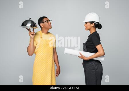 vista laterale dell'ingegnere femmina che tiene il progetto vicino all'uomo in grembiule che tiene isolato sul grigio Foto Stock