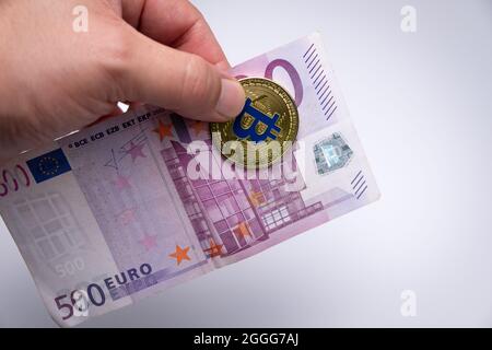 Mano maschile che tiene Golden Bitcoin e una banconota da un euro. Oro moneta BTC di criptovaluta e una banconota da cinquecento euro in mano agli uomini. Foto Stock