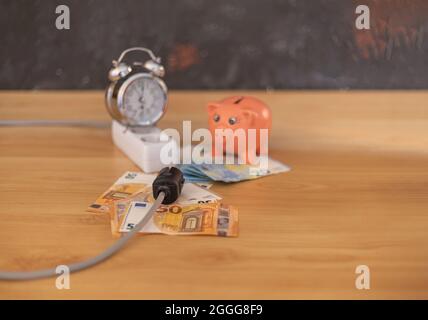 focalizzazione selettiva di un cavo di prolunga di prese elettriche con denaro dal risparmio di una banca piggy e un orologio che indica l'ora Foto Stock