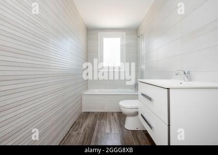 Piccolo bagno bianco con vasca, armadio bianco con cassetti e lavandino sulla destra, con pavimento in ceramica scuro nell'appartamento attico Foto Stock