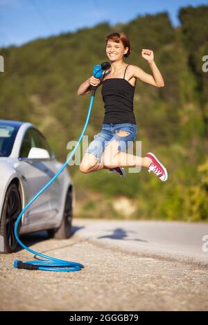 Bella giovane ragazza che salta accanto a un'auto elettrica. Tenendo il cavo di ricarica blu. Foto Stock
