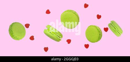 Pezzi di maccheroni di pistacchio verde o di macaroni francesi che si levitano tra piccoli cuori di legno rosso su un morbido sfondo rosa. Fotografia alimentare di levitazione Foto Stock