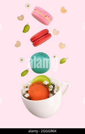Macaroon colorati, piccoli fiori bianchi, foglie di menta verde e cuori di legno che si levitano su una tazza di tè bianco. Isolamento su sfondo rosa tenue. Foto Stock