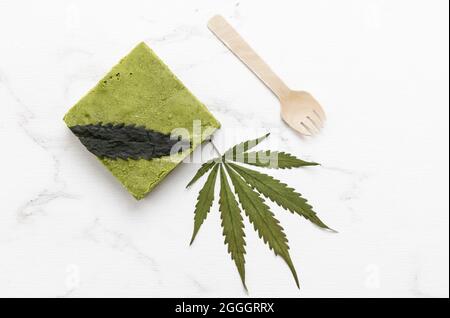 vista di un bar di gustosa torta di cioccolato bianco matcha di cannabis su foglie di cannabis secca e un vassoio di marmo testurizzato Foto Stock